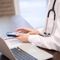 Doctor in a white coat using a calculator next to a laptop on a desk. Doctor in a white coat using a calculator next to a laptop on a desk.