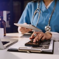A healthcare professional in blue scrubs, with a stethoscope, uses a tablet and calculator at a desk with a laptop and clipboard, conveying diligence.