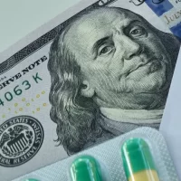 Close-up image showing a partial U.S. hundred-dollar bill with green and yellow capsules in blister packaging. It conveys themes of money and healthcare.