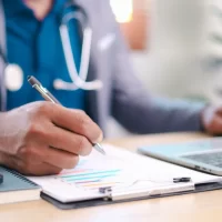 Close-up of professional writing financial report on clipboard at office desk with laptop and smartphone. Concept of analysis, documentation, planning, and consulting work.